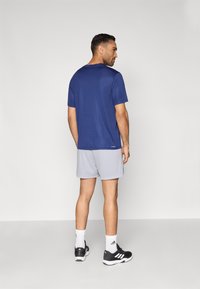 Τζιν μπλε αθλητικό μπλουζάκι με κοντά μανίκια, συνδυασμένο με γκρι σορτς. Το μοντέλο φοράει μαύρα αθλητικά παπούτσια με λευκές λεπτομέρειες και εμφανές λογότυπο.