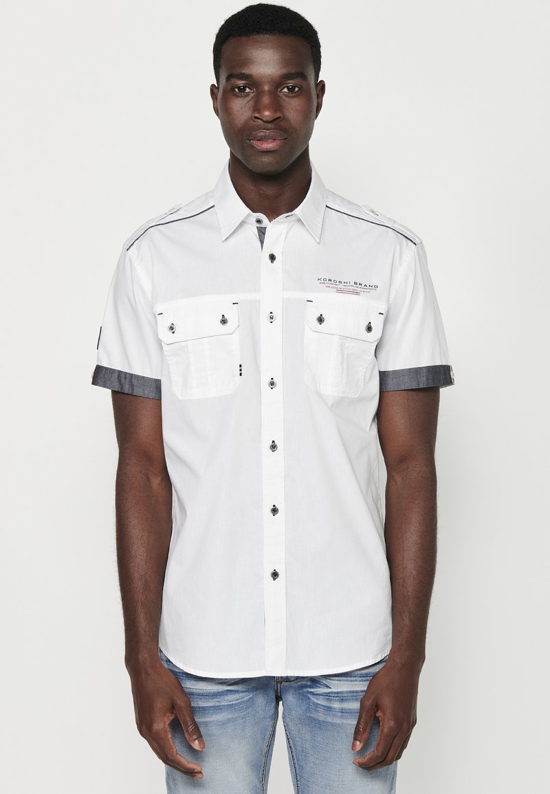 Witte korteklevenshirt met borstzakken, zwarte knopen en denim accenten op de mouwen. Voorzien van een shirtkraag en gestikte details.