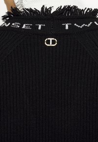 Tissu noir tricoté avec bord à franges, texte blanc "TWINSET" en haut, et petite étiquette logo en métal doré et blanc centrée sous le texte.