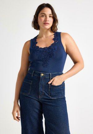 Donna con top smanicato blu navy con dettaglio in pizzo floreale e jeans blu scuro a vita alta, posando su uno sfondo semplice.