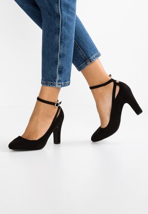 Zwarte suède hoge hakken met een ronde neus en een enkelbandje met een ronde metalen accent; gecombineerd met een cropped blauwe jeans.