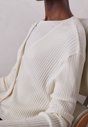 Person wearing a ribbed, cream-colored cardigan over a white top, seated on a light-colored chair against a plain background.