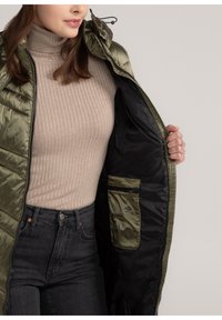 Olijfgroene padded jack met capuchon, open geritst om een zwarte voering en een binnenzak te tonen. Geribbeld beige coltruitje eronder, gecombineerd met donkere jeans.