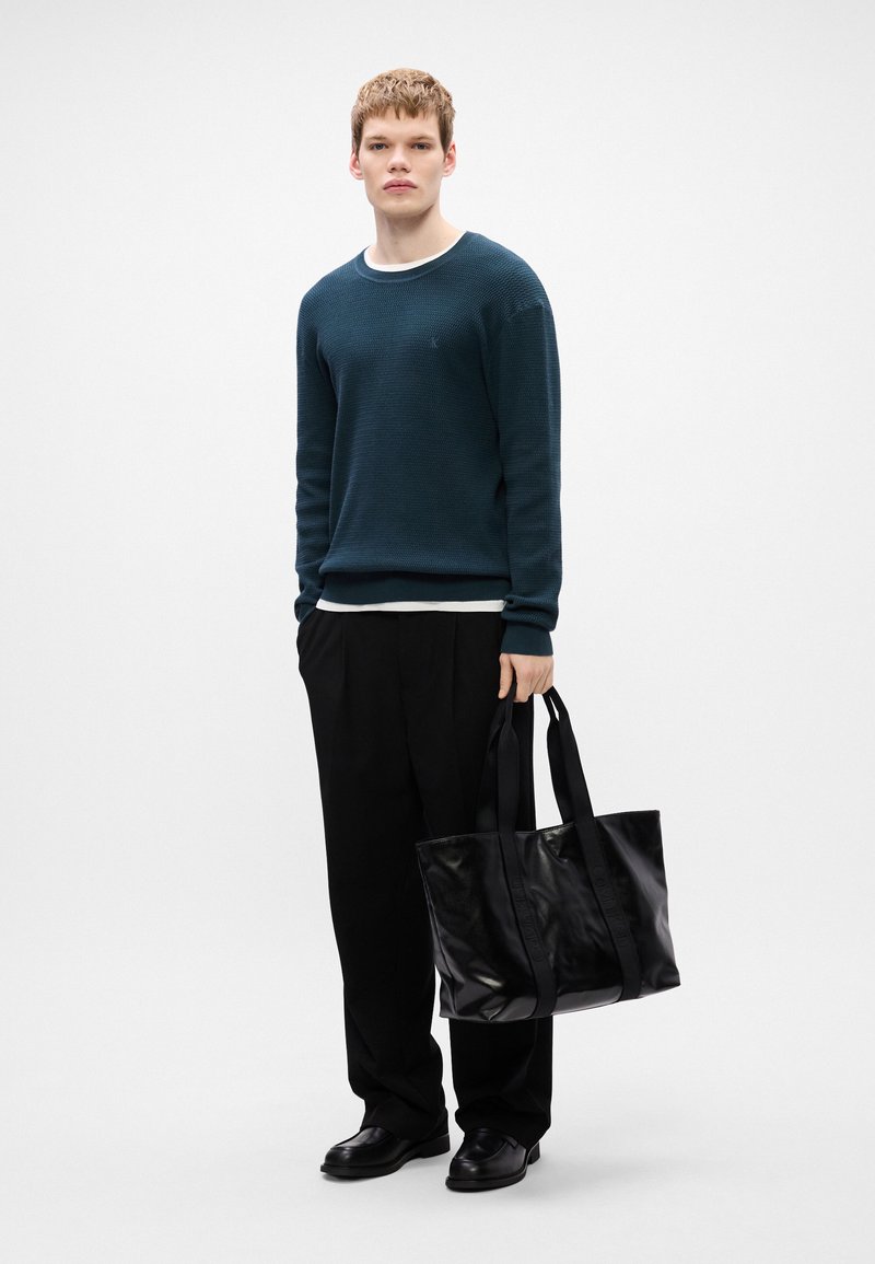 Dark green textured knit sweater over a white crew neck, black trousers, and a shiny black tote bag with shoulder straps. Shoes are black.