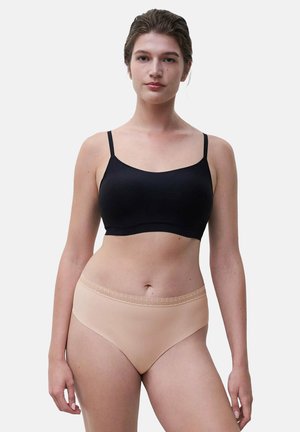 Woman wearing a black cropped camisole and beige high-cut underwear with lace waistband, standing against a plain light background.