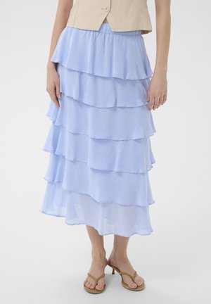Kobieta ubrana w jasnoniebieską, warstwową spódnicę midi z falbanami, beżową bezrękawnikową kamizelkę na guziki oraz brązowe sandały na obcasie na białym tle.