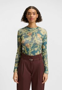Long-sleeve top in sheer, lightweight fabric featuring a floral print in green, yellow, and red tones, paired with high-waisted dark brown trousers.