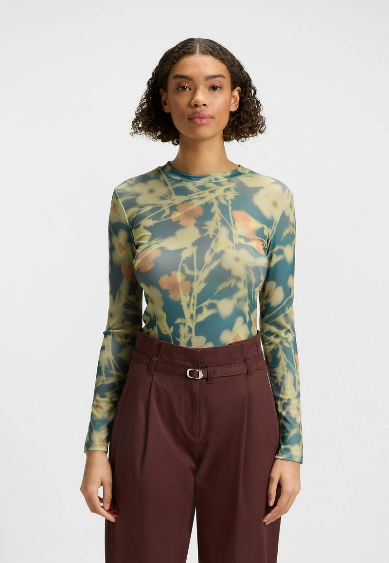 Long-sleeve top in sheer, lightweight fabric featuring a floral print in green, yellow, and red tones, paired with high-waisted dark brown trousers.