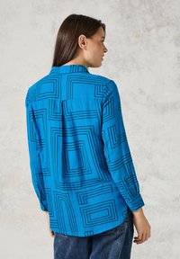 Camisa azul con patrones geométricos de cuadrados negros, de mangas largas y con cuello abotonado. Tejido suave con un acabado ligeramente texturizado.