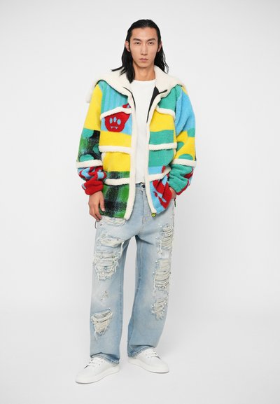 Veste en polaire colorée avec un design en patchwork jaune, bleu, vert et rouge, avec un col et des détails en fausse fourrure blanche. Portée avec un jean déchiré.