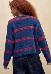 Vrouw met krullend rood haar, gekleed in een fuzzy blauwe trui met rode strepen en donkere spijkerbroek, met de rug naar de camera toegekeerd tegen een neutrale achtergrond.
