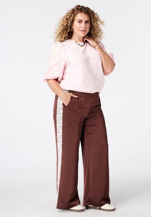 Femme aux cheveux bouclés portant un haut rose à manches bouffantes et un pantalon large marron avec des bandes en dentelle blanche, debout avec une main dans la poche.
