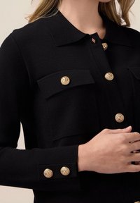 Svart cardigan sydd av texturerat material, med en krage och fyra knappar i guldton framtill och på ärmarna.