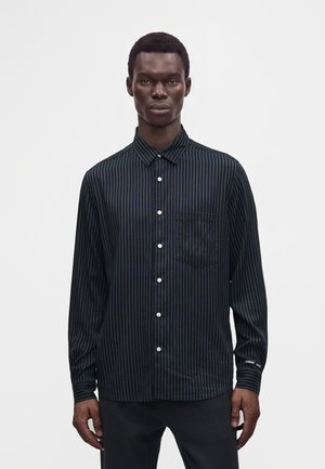 Homme portant une chemise noire à manches longues avec de fines rayures blanches verticales, debout devant un fond blanc uni.