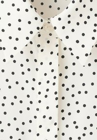 Weißer Stoff mit kleinen schwarzen Tupfen, spitzer Kragen und mittige, mit Knöpfen versehene Blende.