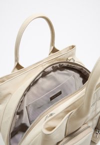 Sac fourre-tout beige en matériau doux, doté de deux poignées, d'une poche intérieure zippée et d'une doublure lisse avec une étiquette.