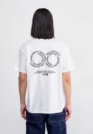 Osoba o ciemnych włosach do ramion nosi białą koszulkę z czarnym okrągłym napisem oraz ciemne spodnie typu cargo, stojąc tyłem do kamery.
