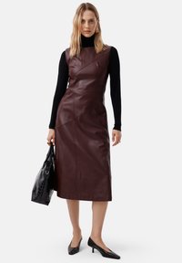 Abito midi in pelle bordeaux con cuciture sulle spalle, pince curvate e tasca laterale. Abbinato a décolleté nere con punta a punta e una borsa a tracolla nera.