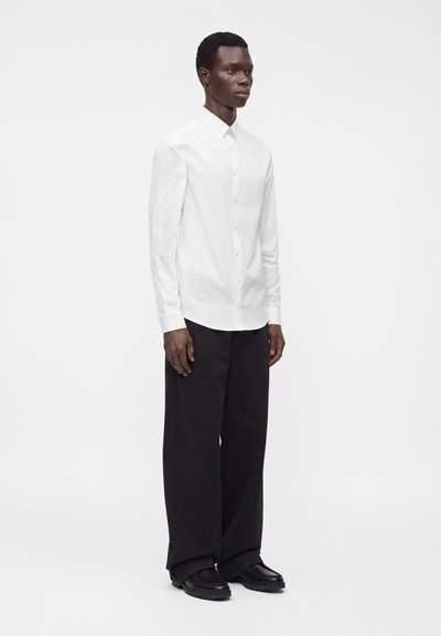 Uomo in piedi che indossa una camicia bianca con bottoni, pantaloni neri a gamba larga e scarpe nere su uno sfondo bianco uniforme.