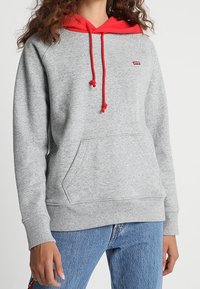 Sweat-shirt gris avec capuche et cordons rouges, doté d'une poche kangourou à l'avant et d'un petit logo. Fabriqué en tissu doux et texturé.
