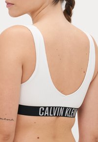 Soutien-gorge de sport blanc à côtes avec une large bande élastique noire portant "CALVIN KLEIN" en texte blanc, affichant un design à dos en scoop.
