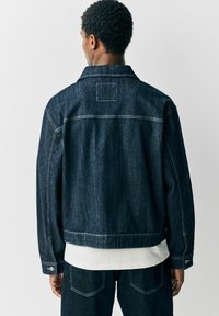 Denim jasje in donker indigo met een klassieke snit, contrasterende stiksels, knoopsluiting bij de mouwen en een achterpatch. Zoom zit onder de taille.