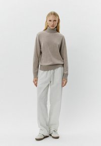 Pulover bej cu guler înalt și margine împletită, combinat cu pantaloni largi cu dungi deschise. Modelul poartă adidași de culoare deschisă. Fundal neutru.