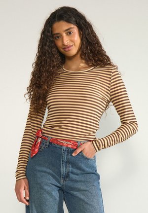 Vrouw met lang krullend haar, gekleed in een bruin-beige gestreept shirt met lange mouwen, blauwe spijkerbroek en een rode bandana-riem, staand met hand in de zak.