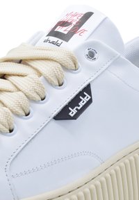 White leather sneaker with cream laces, black logo accents, and textured rubber sole. Features a printed tongue with text.