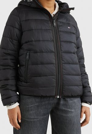 Person iført en sort quiltet jakke med frontlynlås og lynlåslommer over en hvid sweater og mørke jeans.