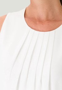 Haut sans manches blanc avec des plis verticaux au niveau du col, texture lisse et col légèrement arrondi.