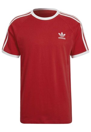 Mężczyzna w czerwonej koszulce Adidas i dopasowanych czerwonych spodniach z białymi paskami i logo na rękawach i klatce piersiowej.