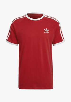 Mies pukeutunut punaiseen Adidas-t-paitaan ja siihen sopiviin punaisiin housuihin, joissa on valkoiset raidat ja logot hihoissa ja rinnassa.