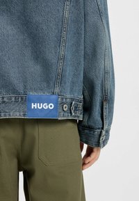 Jeansjacka med en ljusblå tvättning, med en blå etikett med "HUGO"-text och silverfärgad hårdvara. Bärs med olivgröna byxor.