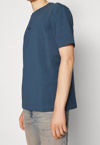 Tmavě modré bavlněné tričko s krátkými rukávy, kulatým výstřihem a jemným logem na hrudi. Měkká textury a uvolněný střih. Jednoduchý design.