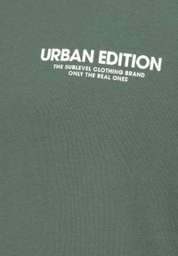 Grüner Baumwollstoff mit dem Text "URBAN EDITION" in fettem Weiß, begleitet von kleinerem Text in derselben Farbe. Insgesamt glatte Textur.