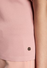 Light pink knitted top with a smooth texture and short sleeves. Features a small metallic logo accent on the lower side.