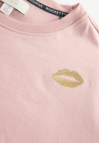 Šviesiai rožinė marškinėlių su minkštu audiniu, priekyje auksinė siuvinėta lūpų dizainas. Kaklo apykaklės detalė su ribomis.