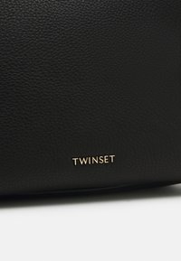 Close-up van zwart gestructureerd leer met kleine gouden metalen letters die "TWINSET" spellen op een witte achtergrond.