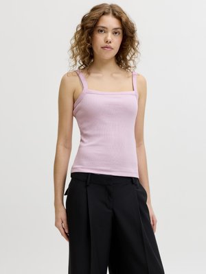 Jeune femme aux cheveux bouclés portant un débardeur rose clair à encolure carrée et un pantalon noir ample, debout devant un fond clair uni.