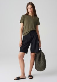 Olive green ribbed t-shirt, black tailored shorts, black flat sandals, and a textured dark green woven tote bag.