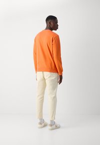 Oranssi huppari, jossa on ribbireunat, beiget slim-fit-housut ja valkoiset sneakersit. Yksinkertainen muotoilu, sileä kangas ja minimaaliset yksityiskohdat.