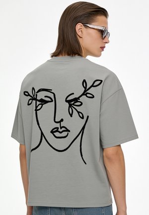 Person trägt ein graues T-Shirt mit schwarzem abstraktem Gesicht- und Blätterdesign auf dem Rücken, kombiniert mit Sonnenbrille und Jeans.