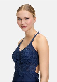 Granatowa sukienka wieczorowa z detalami z koronkowej tkaniny, przezroczystymi elementami oraz koralikowymi akcentami. Posiada regulowane ramiączka oraz dopasowany krój.
