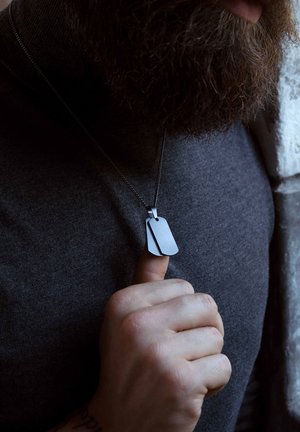 Silver dog tag necklace with two asymmetrical tags, matte finish, on a thin chain. Worn over a dark gray turtleneck. Hand holding necklace.