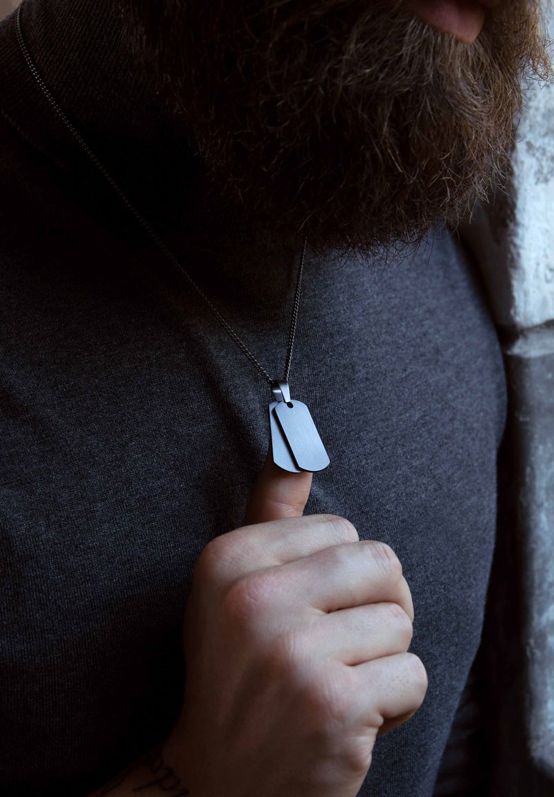 Zilveren hondenpenning ketting met twee asymmetrische tags, matte afwerking, op een dunne ketting. Draagt over een donkergrijze coltrui. Hand houdt de ketting vast.