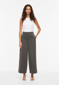 Valkoinen hihaton paita yhdistettynä löysälahkeisiin, tummanharmaisiin housuihin, joissa on sileä pinta; malli seisoo käsineen taskuissa yksivärisen taustan edessä.