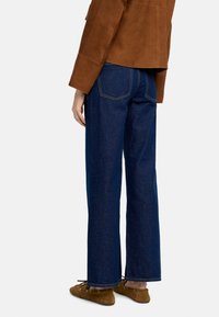 Szerokie, ciemnoniebieskie dżinsy z widocznym przeszyciem i dwoma tylkami. Połączone z brązową zamszową kurtką i brązowym obuwiem.