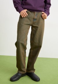 Brown, textured jeans with a relaxed fit and contrast stitching. Paired with black slip-on shoes, worn with a purple hoodie.