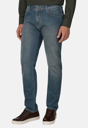 Homme portant un jean droit bleu moyen avec des baskets en cuir marron foncé et une chemise manches longues vert foncé.